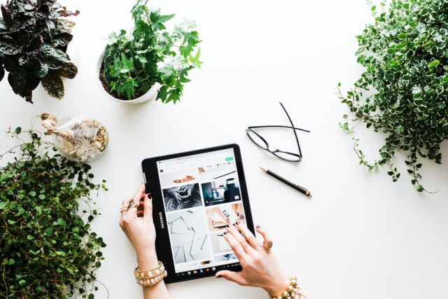 persons hands scrolling on an ipad screen, with a pair of glasses next to them and plants surrounding