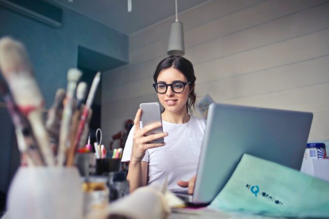 girl on phone and laptop with paintbrushes and creative tools in the back