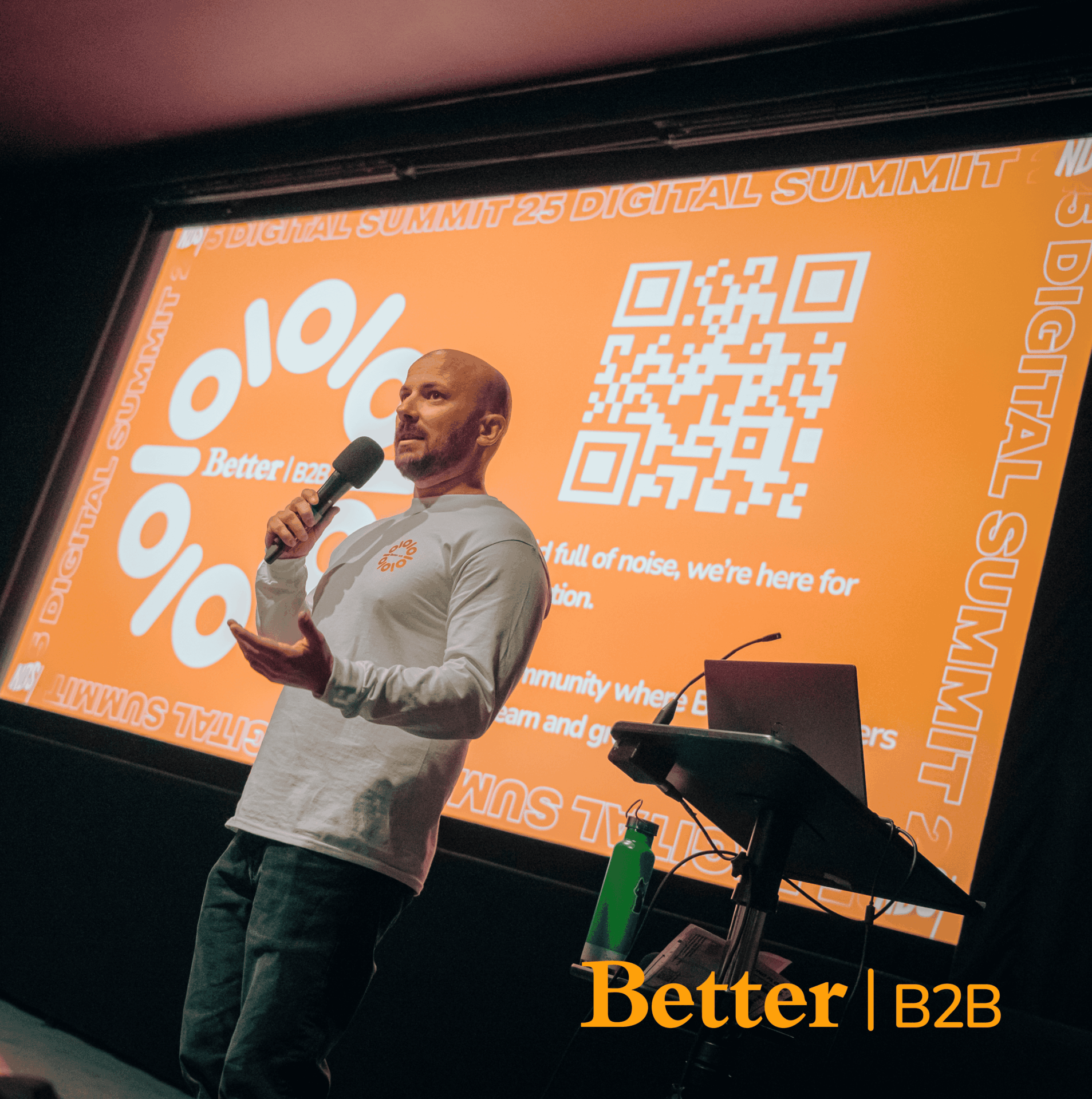 A speaker presenting at the Better B2B Digital Summit, holding a microphone in front of an orange branded slide with a QR code