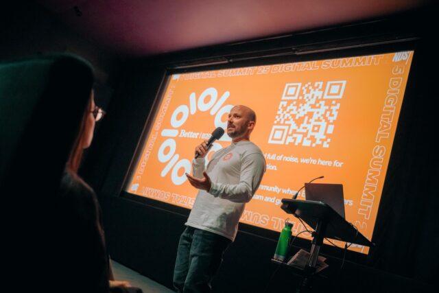 A speaker holding a microphone presents at the NDS Digital Summit 25, with an orange branded slide and QR code displayed behind them